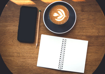 Open blank of note book with latte coffee and mobile phone on wooden table, top view and vintage...