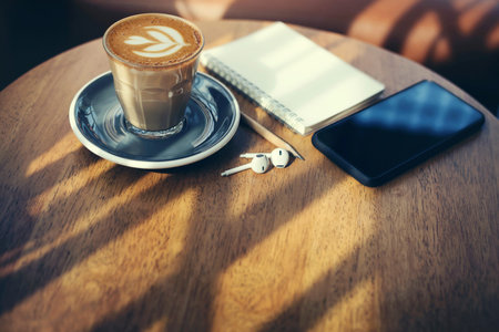 Close-up, latte coffee in old style glass and mobile phone, earphone, note book on wooden table with sunlight, vintage toneの写真素材