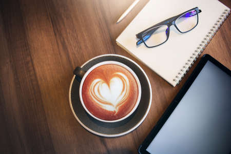 Work from home, desk office with digital tablet, blank notepad, eye glasses, mobile phone and coffee cup with heart shape of latte on wooden table, top viewの写真素材