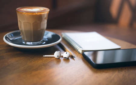 Heart shape of latte coffee in old style glass and mobile phone, note book, wireless headphones on wooden table with vintage sofa and sunlightの写真素材