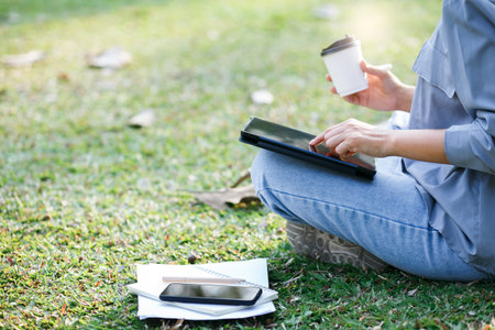Young asian woman with digital tablet sits on the grass in the park. she using tablet online...