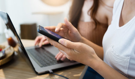 Close up of hand, young asian woman using smart phone working online and her friend using laptop...