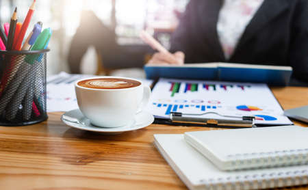 Focus on white cup with latte coffee on desk office and blur background young asian businesswoman working with paperwork and chart on desk officeの写真素材