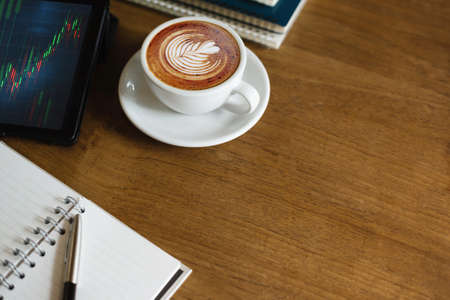 Close up view, Latte coffee in white cup on wooden table with notebook, pen and tablet computer have graph of investment on screen, concept business people relax and working part time in cafeの写真素材