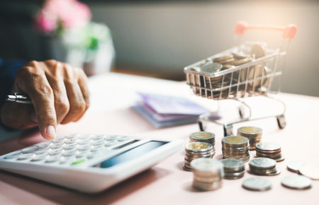 Hand of businessman using calculator to calculate money from savings with stack of coins and coin in small supermarket cart at backgroung. concept of saving for the future and family , close-up viewの写真素材
