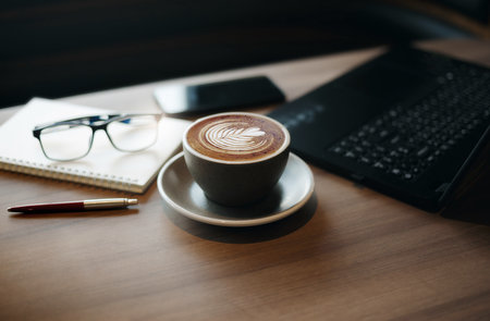 Close-up view, white cup of coffee with computer laptop, notebook, pen and eye glasses on wooden table in cafe, People resting after work in a calm cafeの写真素材