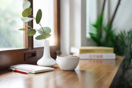 Close-up view, white coffee cup on wooden counter and note book, pen with ceramic vase and small tree by window, coffee break and relax after work in cafe with a reading book in the backgroundの写真素材