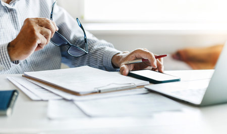 Asian businessman using mobile phone with laptop reviewing document reports at office workplace....
