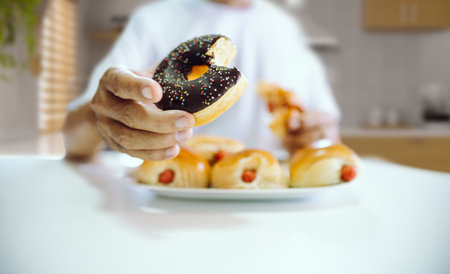 Close-up shot, a man sitting in kitchen holding donut with chocolate on top to eat and many bread with hot dog inside on plateの写真素材