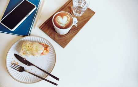 Top view, latte coffee with water in glass and bread for breakfast on white table with mobile phone and notebook before start workingの写真素材