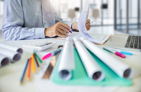 Asian architect in his office review contracts about building designs with blueprint rolls and...