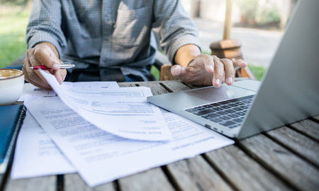 Asian man using laptop working outdoor on old wooden table in plublic park checking documents and...