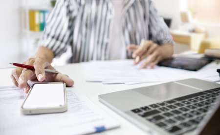 Close-up of hand using mobile phone with documents and working on laptop to searching for...