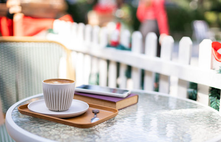 Coffee cup with book for read and mobile phone on glass table next to white fence and sunlight in...