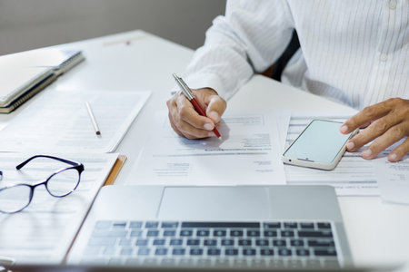 asian businessman using mobile phone with laptop working online at home checking financial documents...