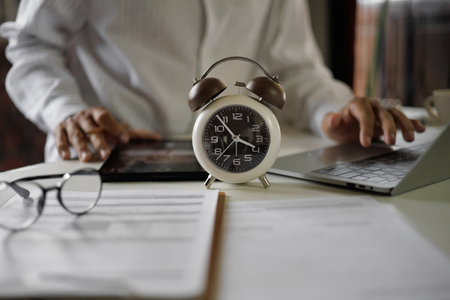 selective focus on alarm clock, asian businessman using laptop and tablet working on table before work time end in a day,  time management concept, routine work, office hour, blur backgroundの写真素材