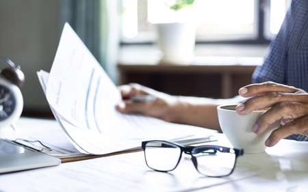 asian businessman working at home reading business document he taking a break during work in afternoon with coffee drink, select focus on hand holding coffee cupの写真素材