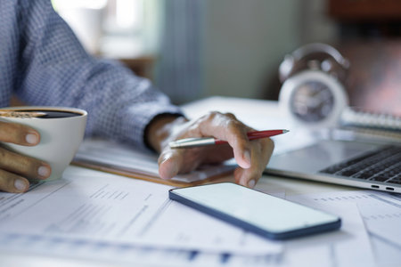 man hand using mobile working with laptop on desk and holding coffee cup during workの写真素材