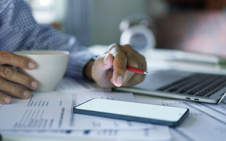 man hand using mobile working with laptop on desk and holding coffee cup during workの写真素材