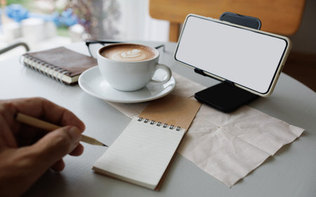 smart phone on stand with latte coffee, note book, diary and man hand holding pencile to taking noteの写真素材