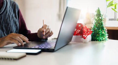 close up shot, asian man using laptop working and decoration christmas background with red hat, christmas tree on desk, working on christmas holiday conceptの写真素材
