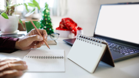 close up shot, hand holding pencil taking note with blank calendar working with laptop, christmas tree for decoration on desk, christmas holidays conceptの写真素材