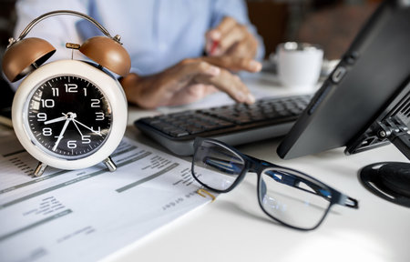 selective focus at alarm clock, asian businessman working with laptop and document on table in the morning, time management concept, routine work, close up shotの写真素材
