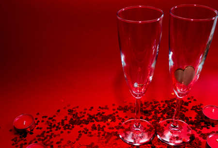 Glasses of champagne, red candles, red confetti in the shape of a heart on a red backgroundの写真素材