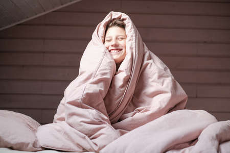 Boy teen sitting the bedroom under the pink blanket over head. Smiling face in the morningの写真素材