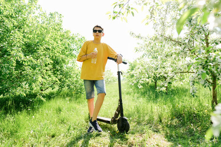 Teen boy drinks water from plastic bottle, standing with scooter at park.の写真素材