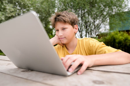 Teen boy with a laptop sits in a park. School boy is typing on computer outdoors. Young boy studying...