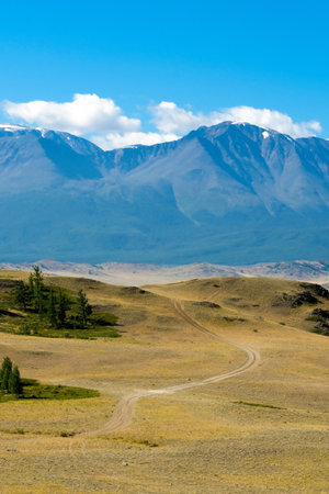 Scenic view from sunlight grassy hill with road to high mountain range. Beautiful sunny mountain landscape with large snow peak mountains. Road trip to Altai.の写真素材