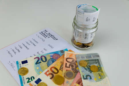 Monthly budget notebook next to banknotes, coins and jar full of rolled up receipts. Increase in tariffs and prices.の写真素材