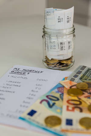 Monthly budget notebook next to banknotes, coins and jar full of rolled up receipts. Increase in tariffs and prices.の写真素材