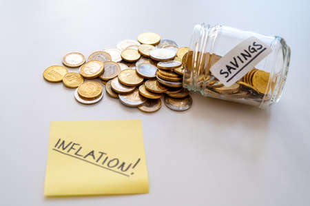 Piggy bank, labeled with word "savings", with coins and yellow note with text "inflaction", on white surface. Rising tariffs and prices, and inflation.の写真素材