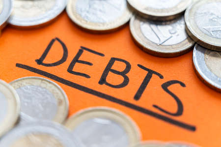 Text "debts", written in black on an orange surface, with coins around it. Rising inflation, debt and economic crisis.の写真素材