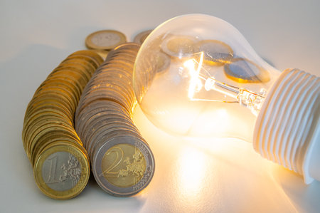 Light bulb lit, with row of coins next to it. Trends in electricity tariffs, energy dependence, energy supplies.の写真素材