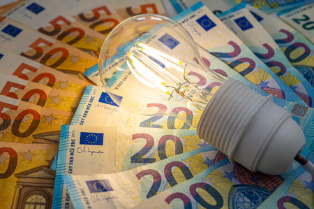 Light bulb lit, above euro banknotes. Electricity costs and tariffs.