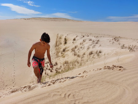 Teenager climbs up a sand ramp in a large area of dunes with a blue sky and few clouds in the background on a sunny day.の写真素材