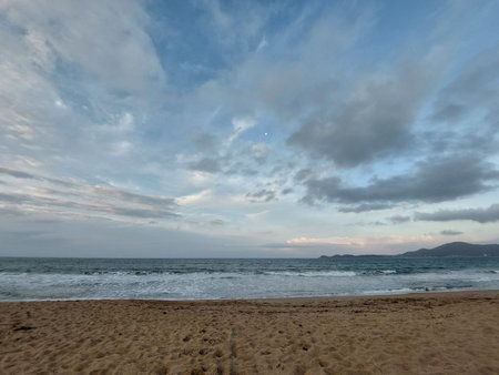 At dusk, on the shore of the beach, gentle waves break, forming lacy foams, and between clouds of soft colors, the moon appears discreetly.の写真素材