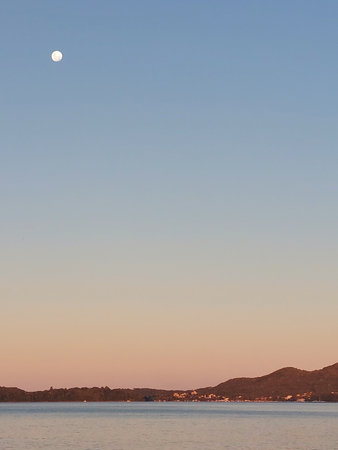At dusk, by the sea, the full moon rises radiantly, leaving the sky in gradient tones and accentuating the contours of the mountains on the horizon.の写真素材