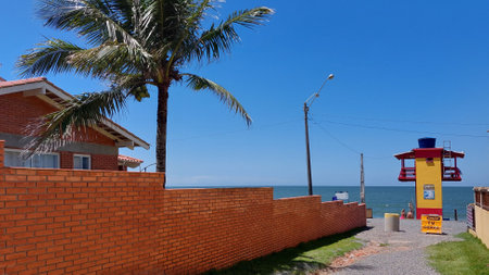 Path to the beach next to the ceramic brick wall passing by the yellow lifeguard tower under the clear blue summer sky.の写真素材
