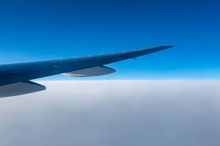 Looking out of an aircraft window at blue sky above smooth white clouds, with the aircraft wing visibleの写真素材