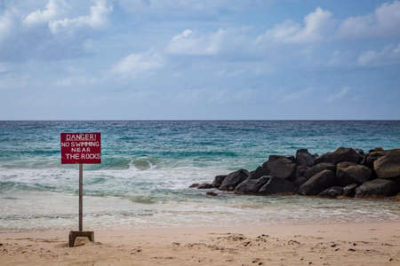 A beach on the island of Barbados with a warning sign near rocksの写真素材
