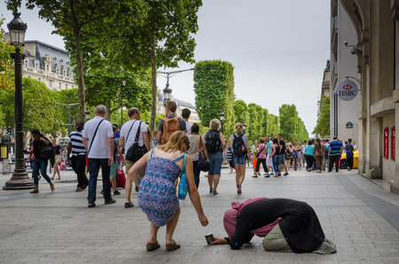 Paris, France, August 8, 2015 - A tourist places coins in the cup of a woman kneeling as she begs for alms on the sidewalk of the Champs-Elysees known for its luxury shops. Crowds pass by without paying attention to the kneeling woman.のeditorial素材