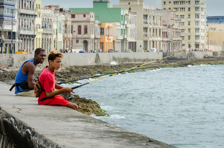 Havana, Cuba, June 20, 2016 - Boy and young man fish from the seawall of the Malecon with historic homes of the city behind them.のeditorial素材