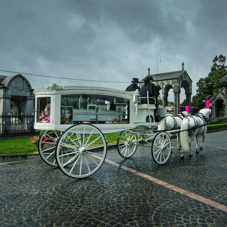 Dublin, Ireland, August 23, 2018 - A white vintage hearse carrying a white coffin and drawn by two white horses with pink plumes enters the Glasnevin Cemetery with dark rain clouds hovering overhead.のeditorial素材