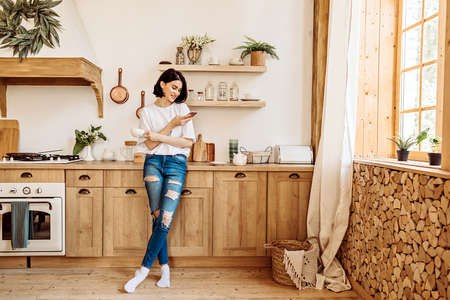 Lifestyle portrait of a happy young girl browsing the news on the phone over a cup of tea or coffee in the kitchen early in the morningの写真素材