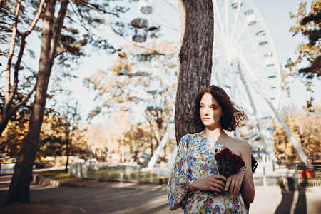 Lifestyle portrait of young woman with bouquet of wildflowers in august sunlight against background of ferris wheelの写真素材