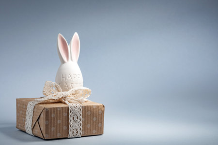 A cheerful Easter bunny figurine sits atop a wrapped gift box, adorned with a lace ribbon, against a soft light blue backdrop. It captures the spirit of the holiday.の写真素材